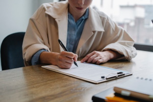 Person i beige jacka sitter vid ett bord och fyller i ett formulär.
